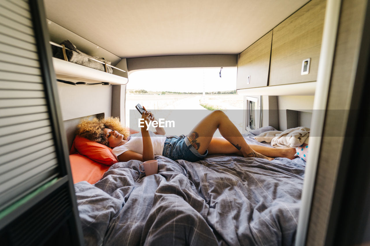 Anonymous woman with afro hairstyle lying upside upon bed in trailer and browsing mobile phone on sunny summer day