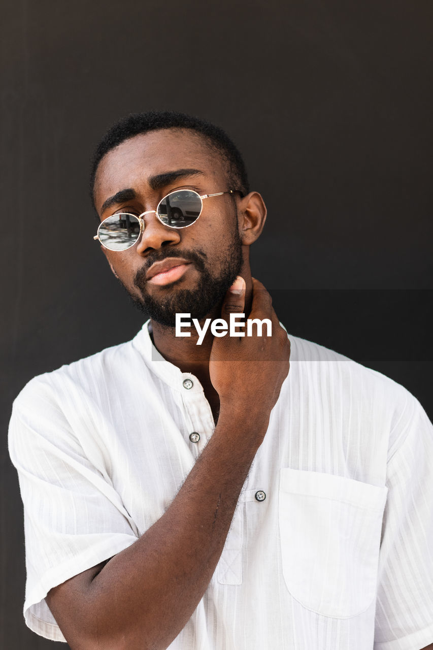 Confident african american male wearing stylish sunglasses and white t shirt standing on street while looking at camera and touching neck