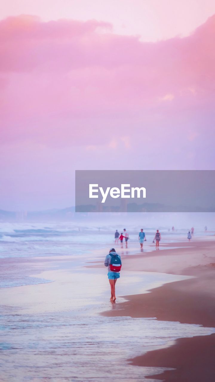 REAR VIEW OF PEOPLE WALKING ON BEACH AT SUNSET