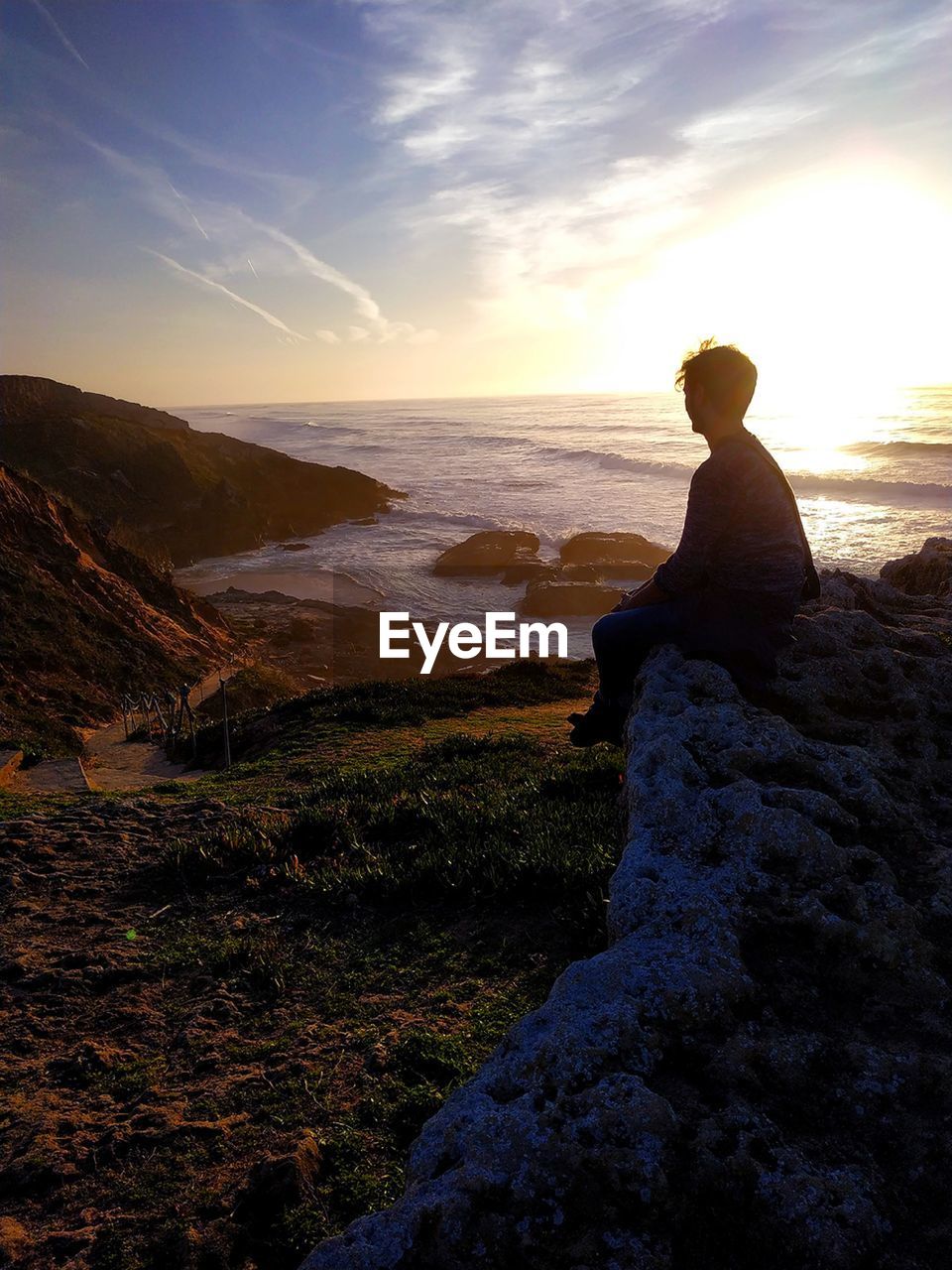 MAN SITTING ON ROCK AT SEA SHORE AGAINST SKY