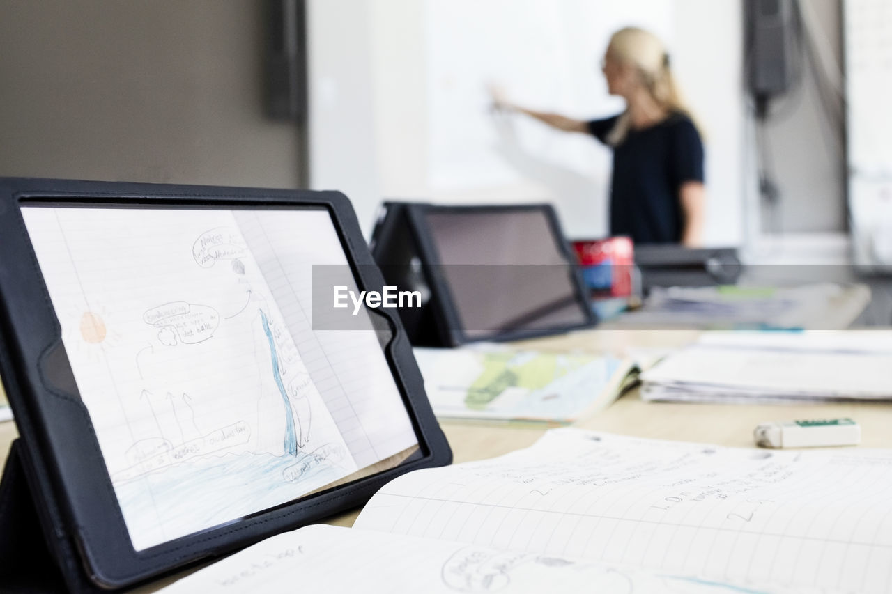 Close-up of digital tablet on desk with teacher explaining in background