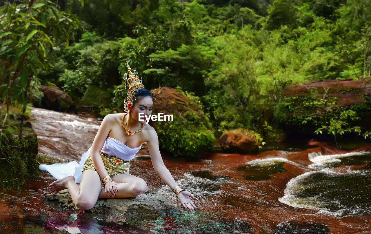 Full length of woman in traditional clothing sitting by stream in forest