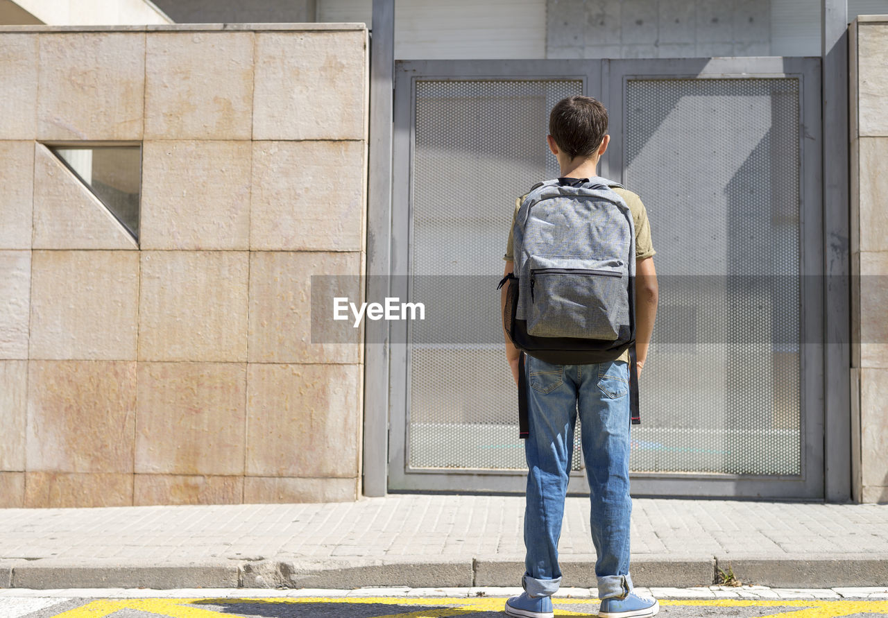 Rear view of boy with backpack standing against metal door during sunny day