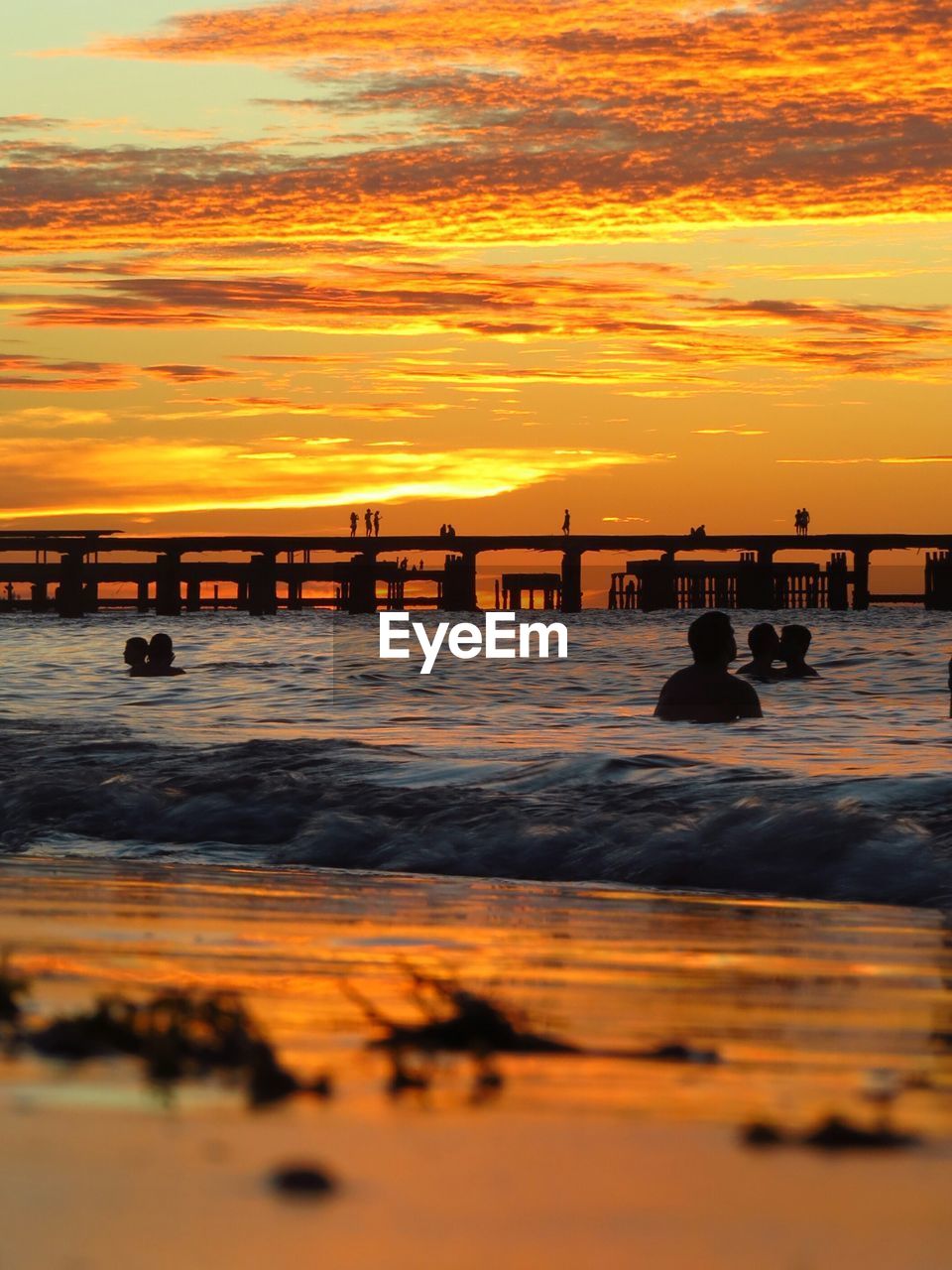 SILHOUETTE PEOPLE SITTING ON SHORE AGAINST ORANGE SKY