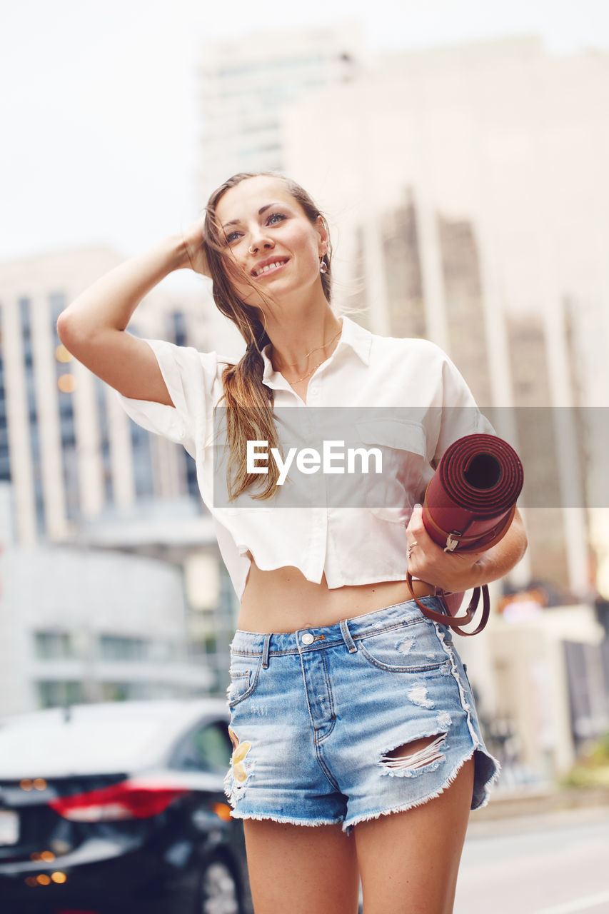 Low angle view of beautiful woman holding exercise mat while standing in city