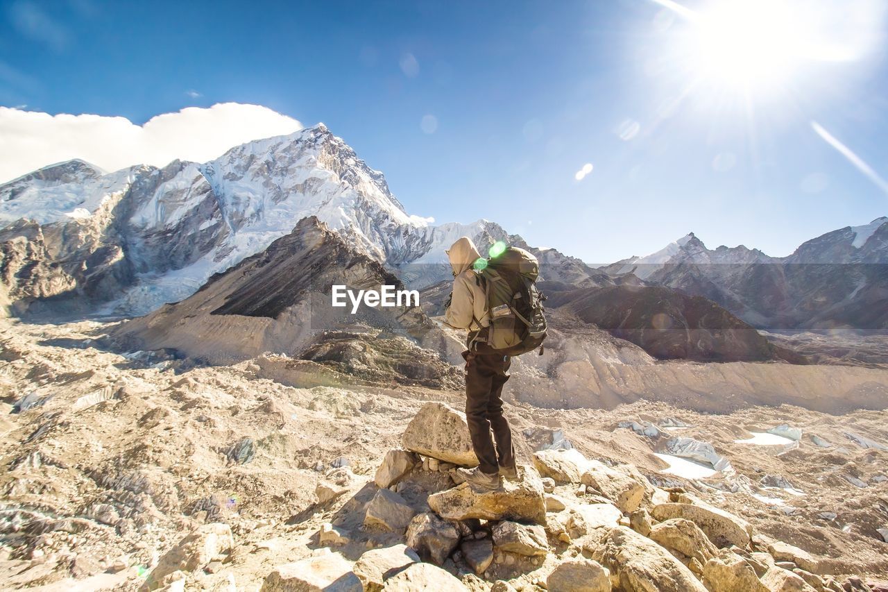 Side view of backpacker standing on rock by mountains against sky during sunny day