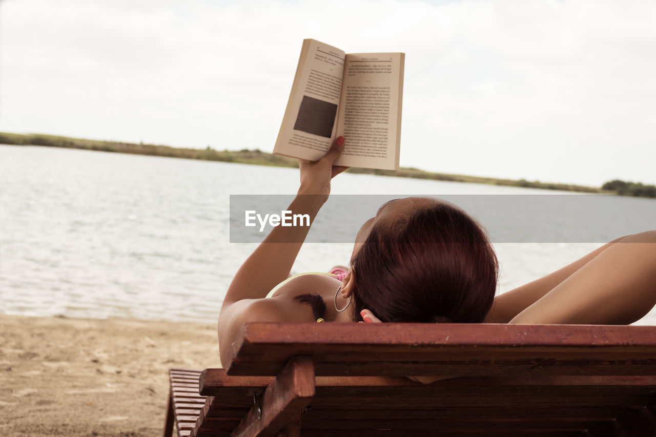 Woman reading book while lying on bench at beach against cloudy sky