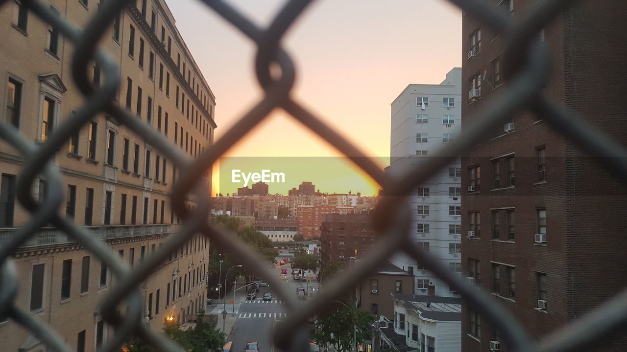 CLOSE-UP OF CITYSCAPE SEEN THROUGH CHAINLINK FENCE DURING SUNSET