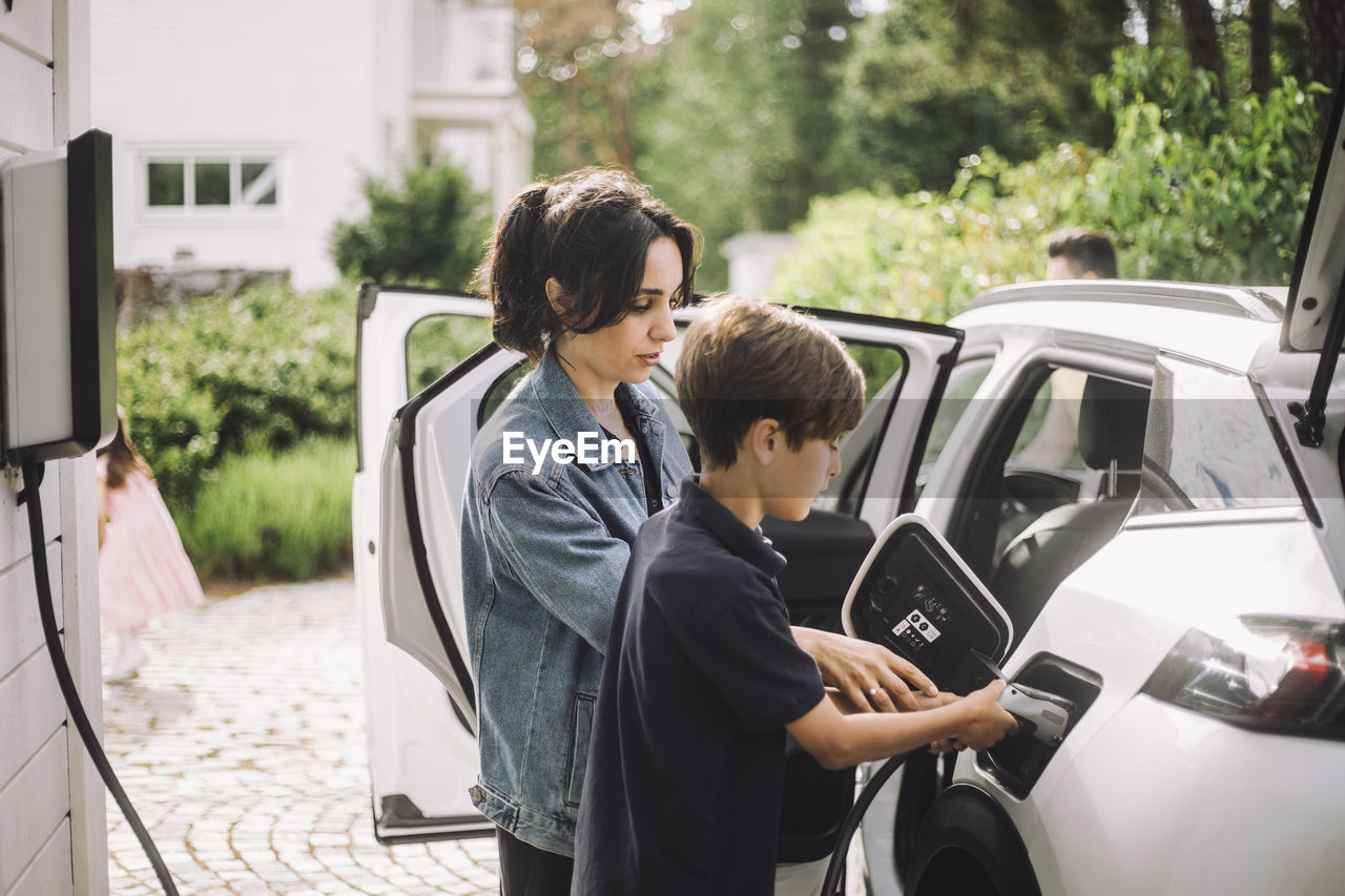 Side view of mother and son charging electric car outside house