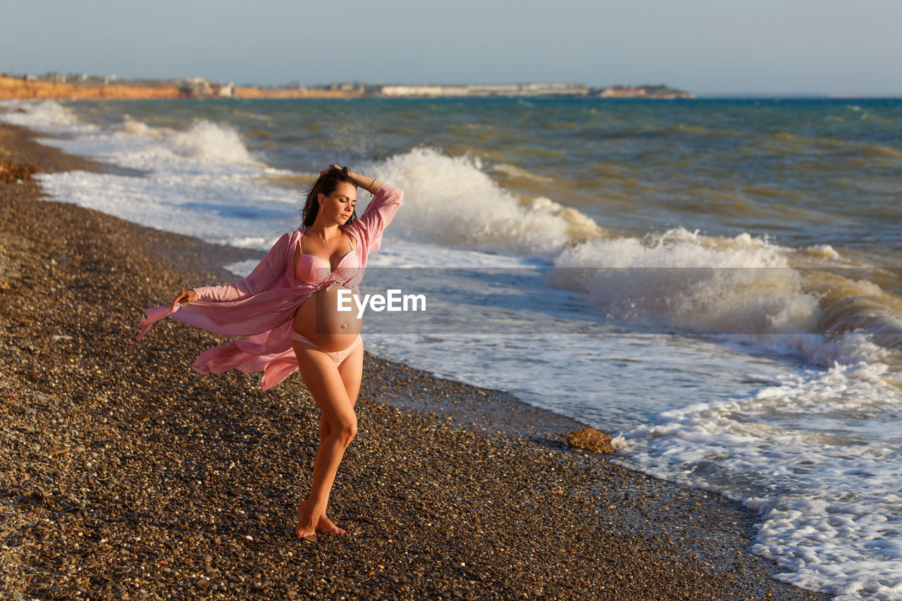 Beautiful pregnant woman standing on beach in light dress in the wind. pleasure