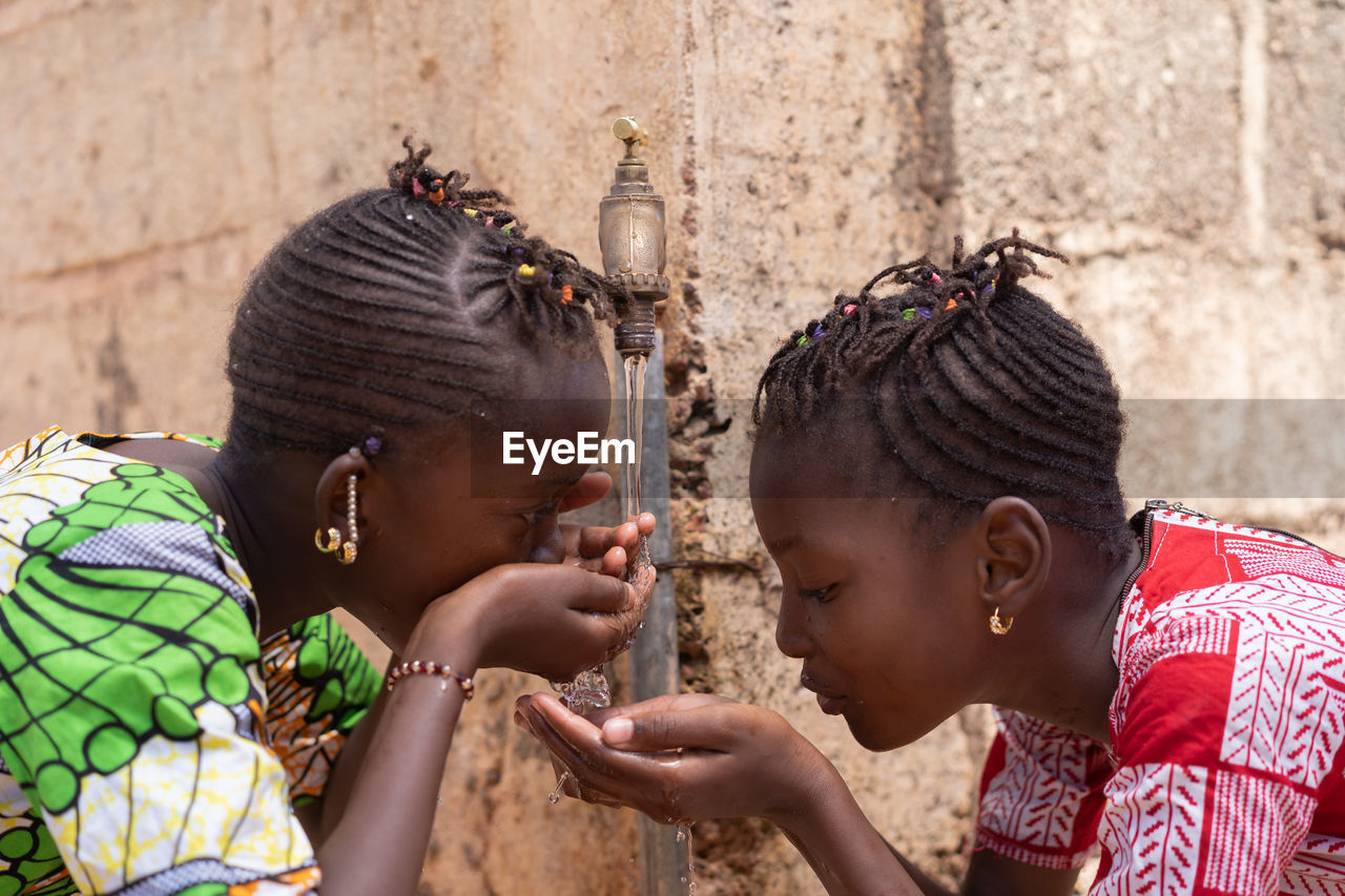 Girls drinking water from faucet against wall