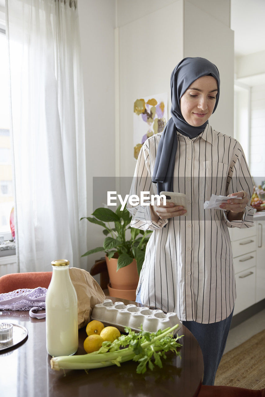Woman in headscarf unpacking groceries at home and checking receipt