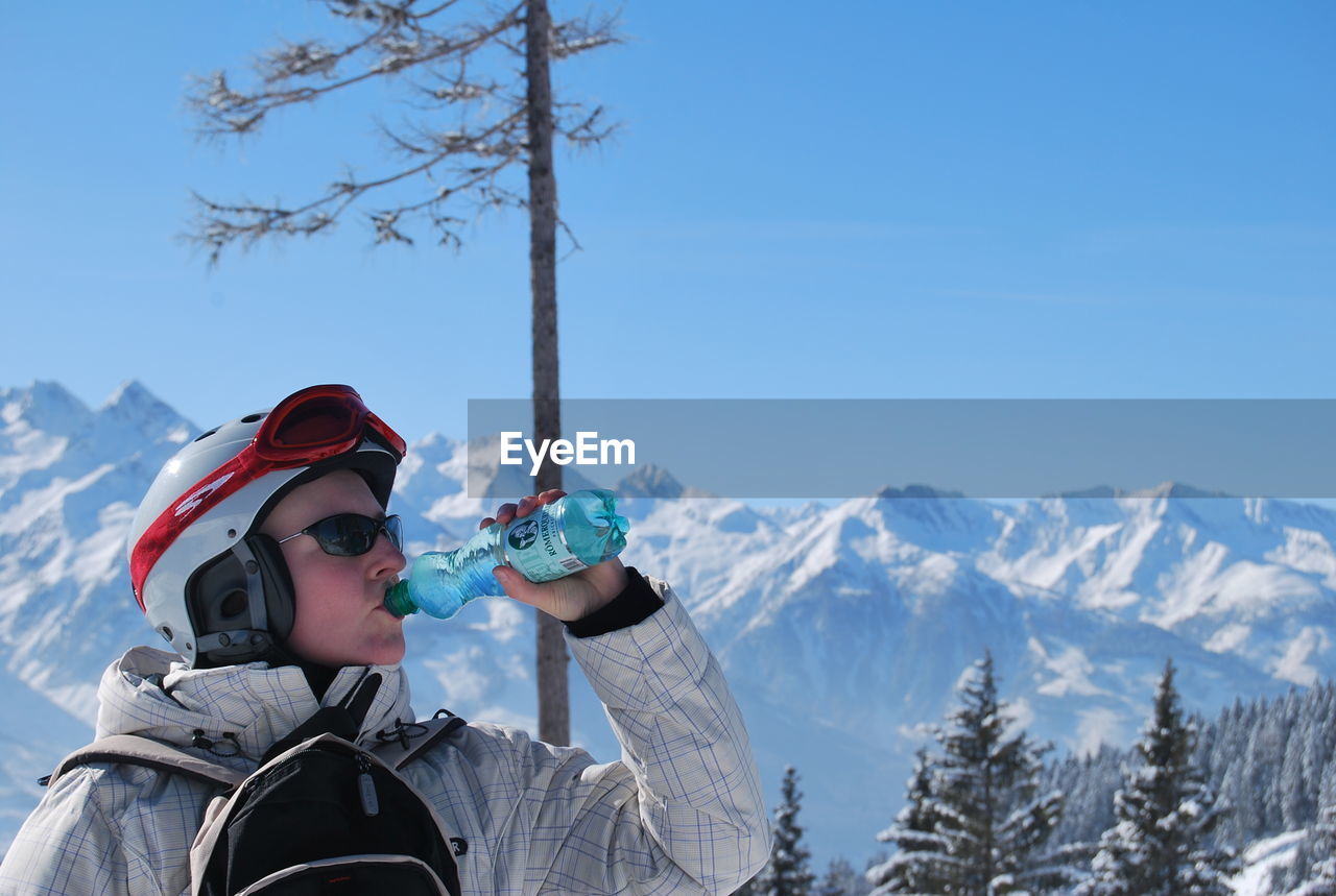 Woman wearing skiing helmet drinking water against snowcapped mountain