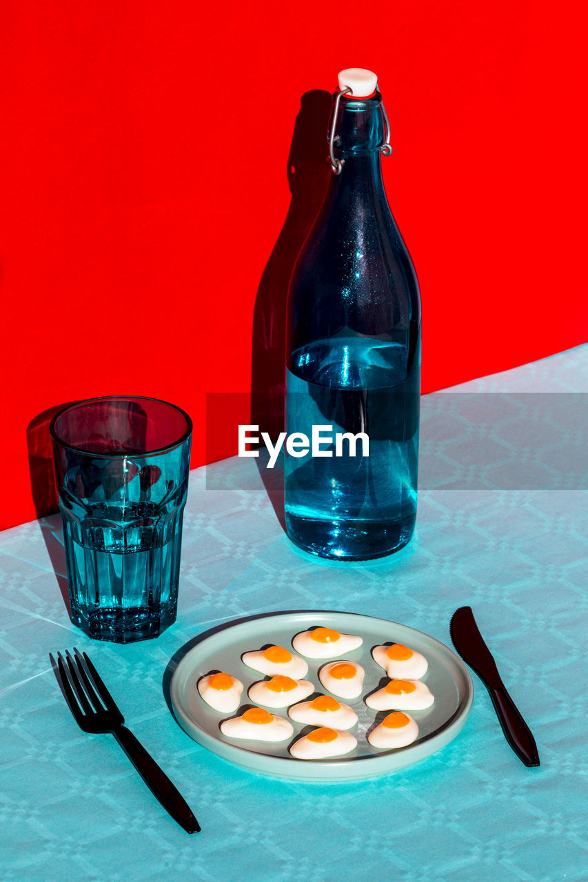 From above of composition of bottle of water with glass opposite plate with halved boiled eggs and fork and knife placed on blue surface against red background