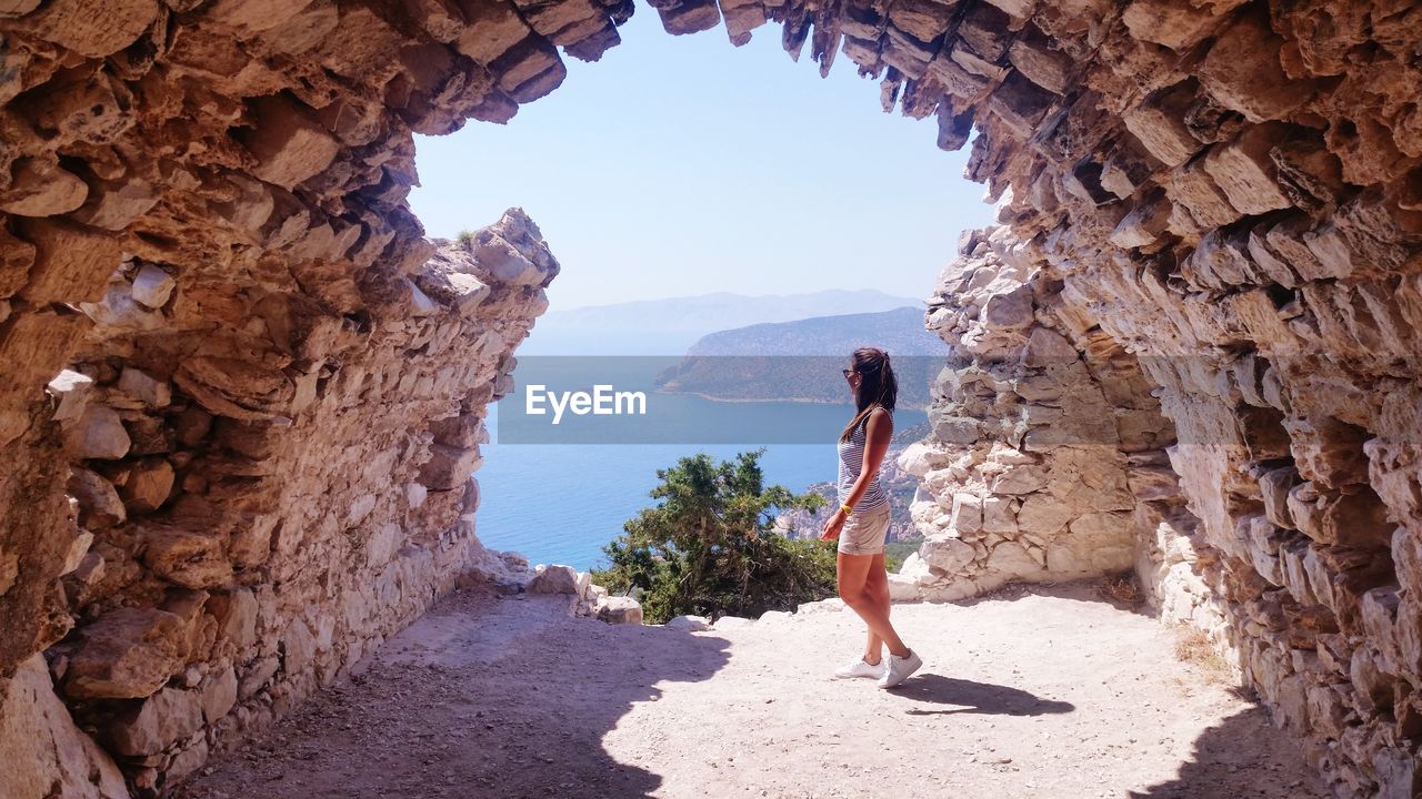 Side view of young woman standing in cave against sky during sunny day