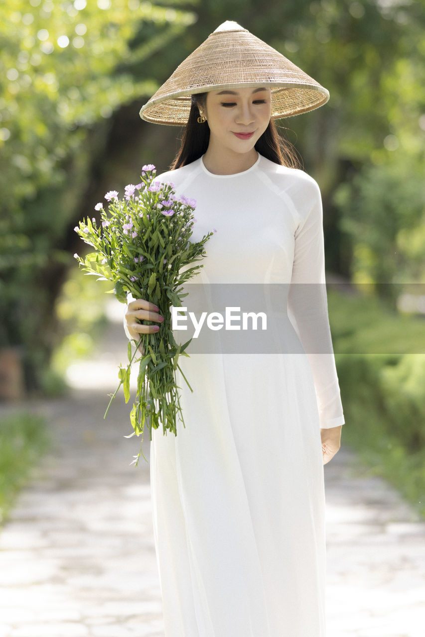 clothing, women, adult, plant, one person, fashion, young adult, nature, flower, gown, wedding dress, female, bride, dress, hat, wedding, flowering plant, bridal clothing, standing, celebration, summer, bouquet, flower arrangement, newlywed, portrait, outdoors, elegance, white, happiness, day, emotion, event, fashion accessory, smiling, holding, spring, person, life events, sun hat, front view, tree, focus on foreground, green, beauty in nature, lifestyles, looking, sunlight, formal wear, brown hair, yellow, glamour, looking at camera, veil, positive emotion