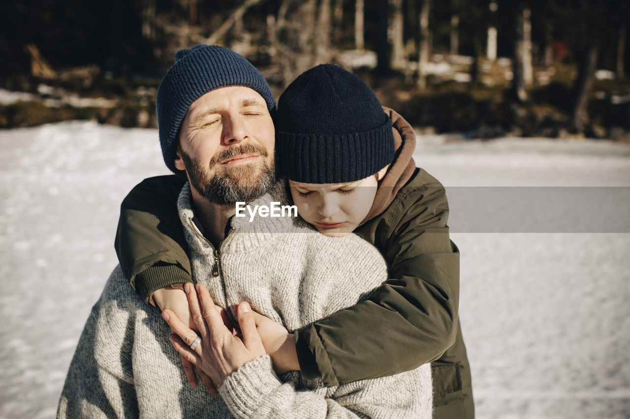 Boy in warm clothing hugging father with eyes closed during sunny day in winter