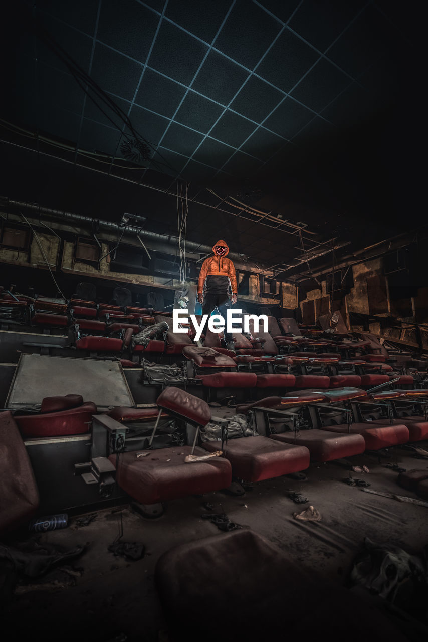 Digital composite image of man wearing mask while standing on broken chair in abandoned movie theater