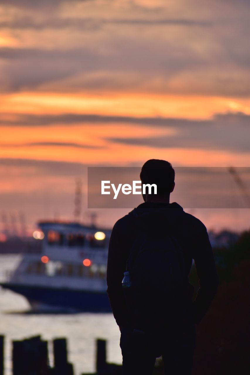 Silhouette man standing at harbor against orange sky