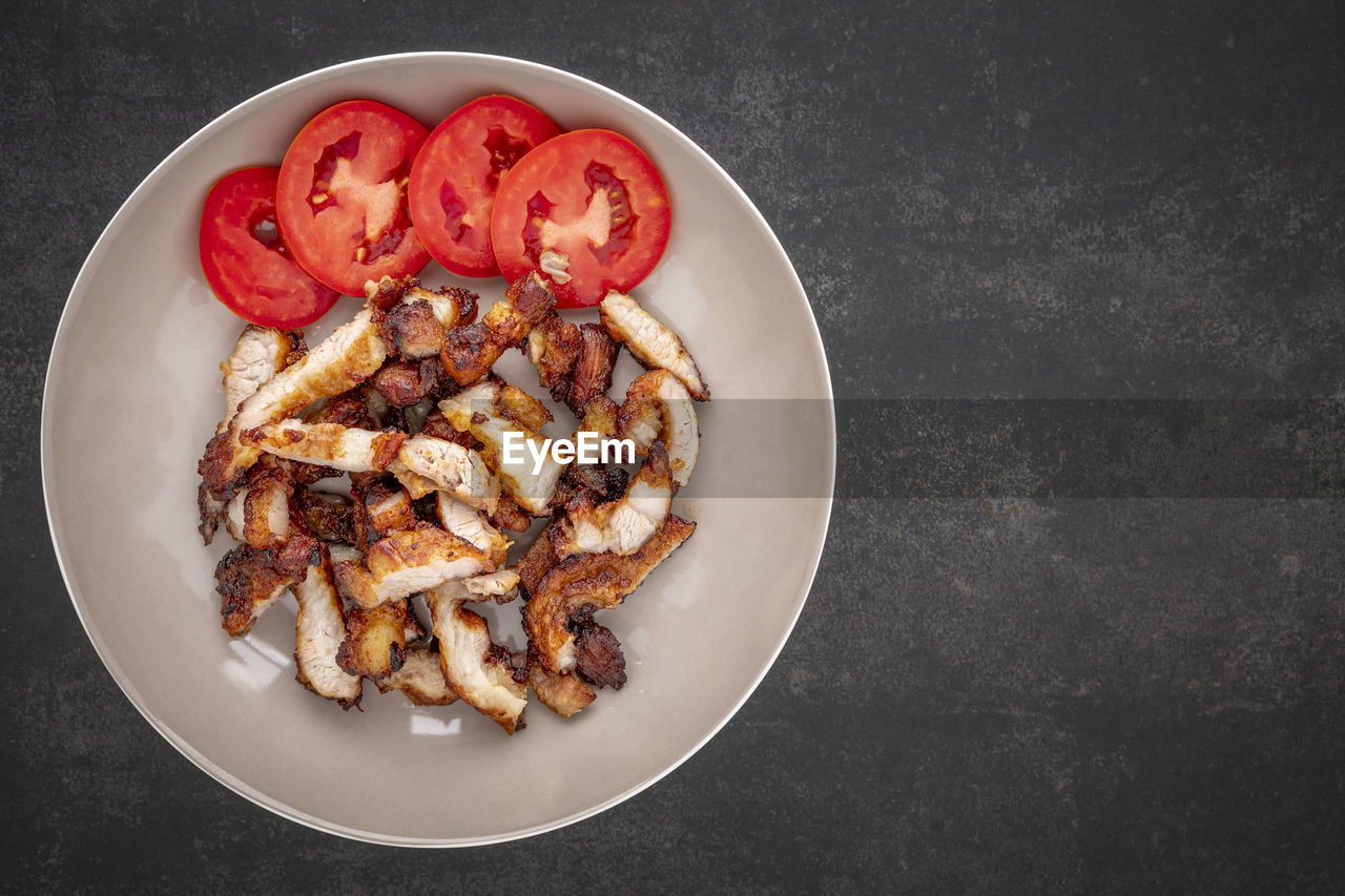 Sliced fried pork with tomato in brown ceramic plate on dark tone texture background, top view