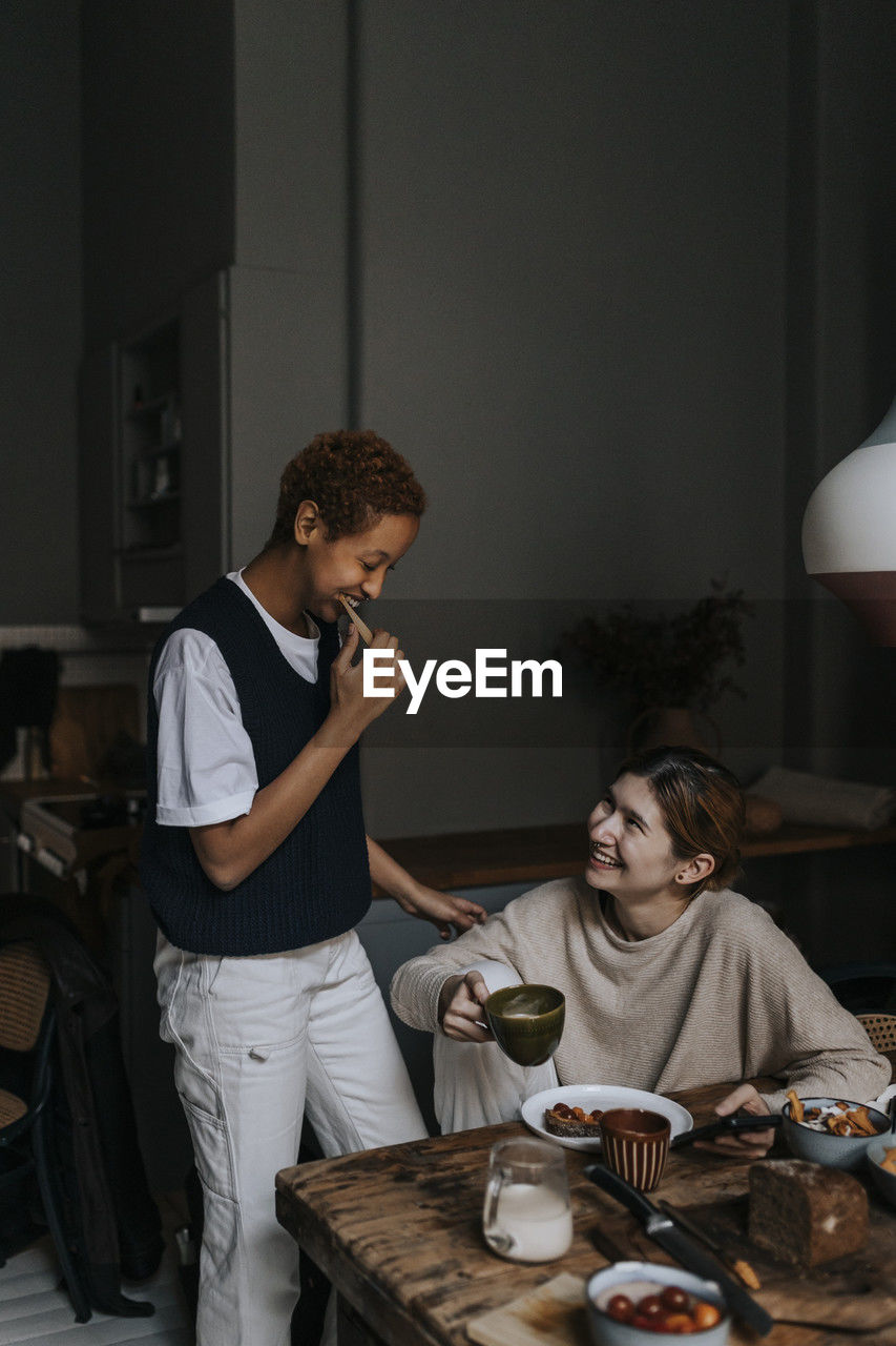 Smiling non-binary person brushing teeth by friend having breakfast at home