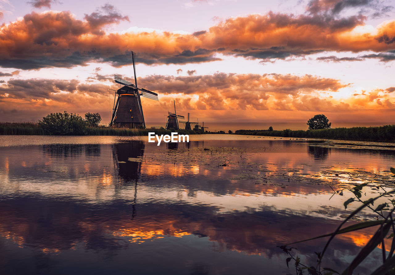 Traditional windmill by lake against sky during sunset