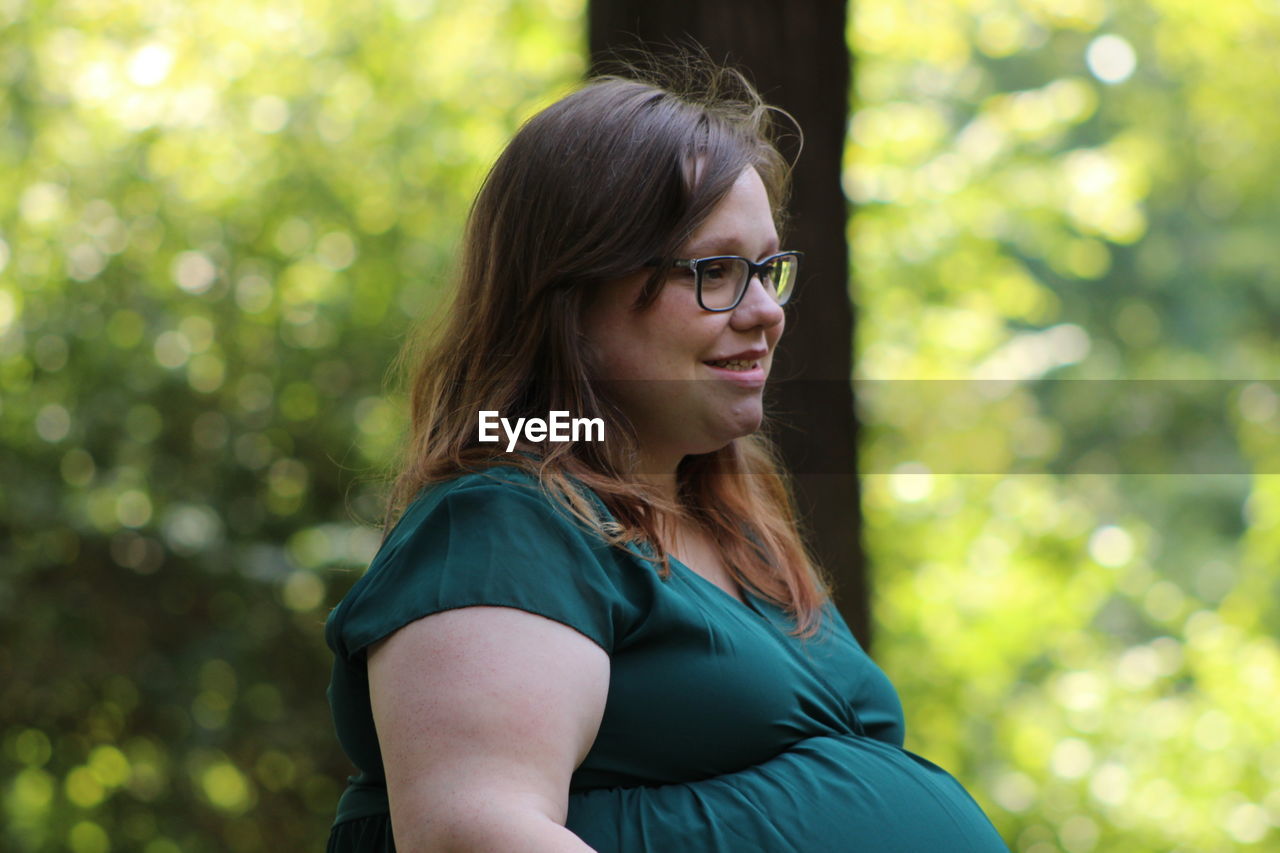 Smiling pregnant woman looking away while standing against trees