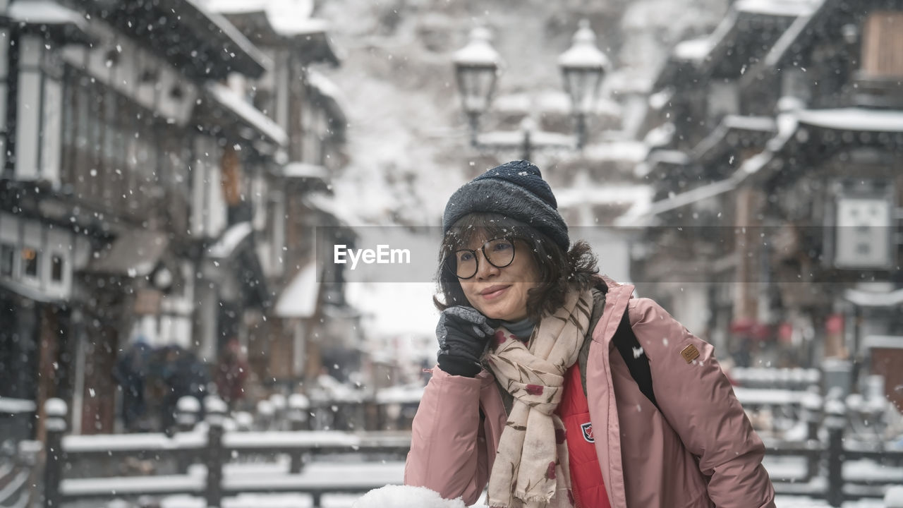 winter, cold temperature, warm clothing, clothing, snow, adult, city, one person, architecture, snowing, hat, women, scarf, young adult, coat, city life, lifestyles, nature, knit hat, smiling, building exterior, portrait, leisure activity, looking, happiness, street, winter coat, front view, female, jacket, waist up, emotion, outdoors, day, city street, human face, focus on foreground, enjoyment, standing, storm, snowflake, frozen, built structure, glove, cheerful, road, person