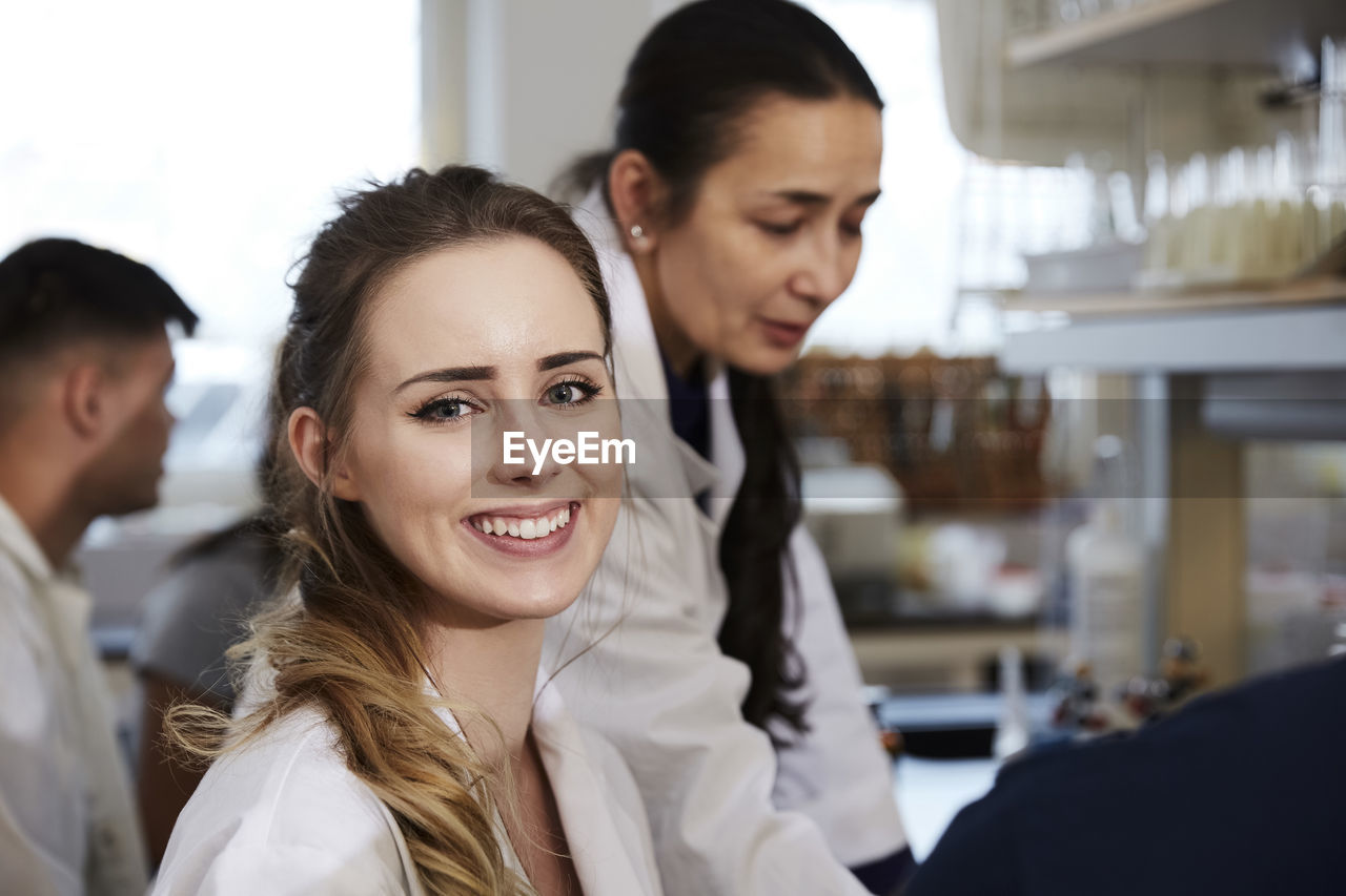 Portrait of smiling young female student sitting against teacher friends in chemistry laboratory