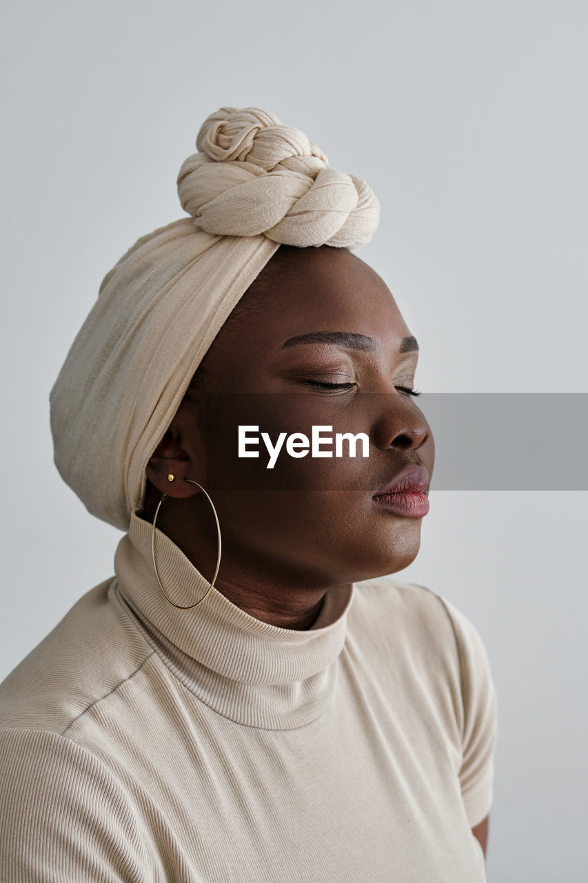 Gorgeous young african female model in stylish traditional turban standing against white background with closed eyes