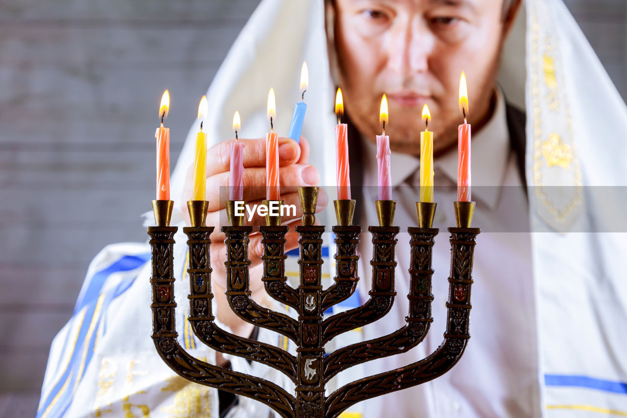 Mature man in jewish prayer shawl burning candles on hanukkah menorah