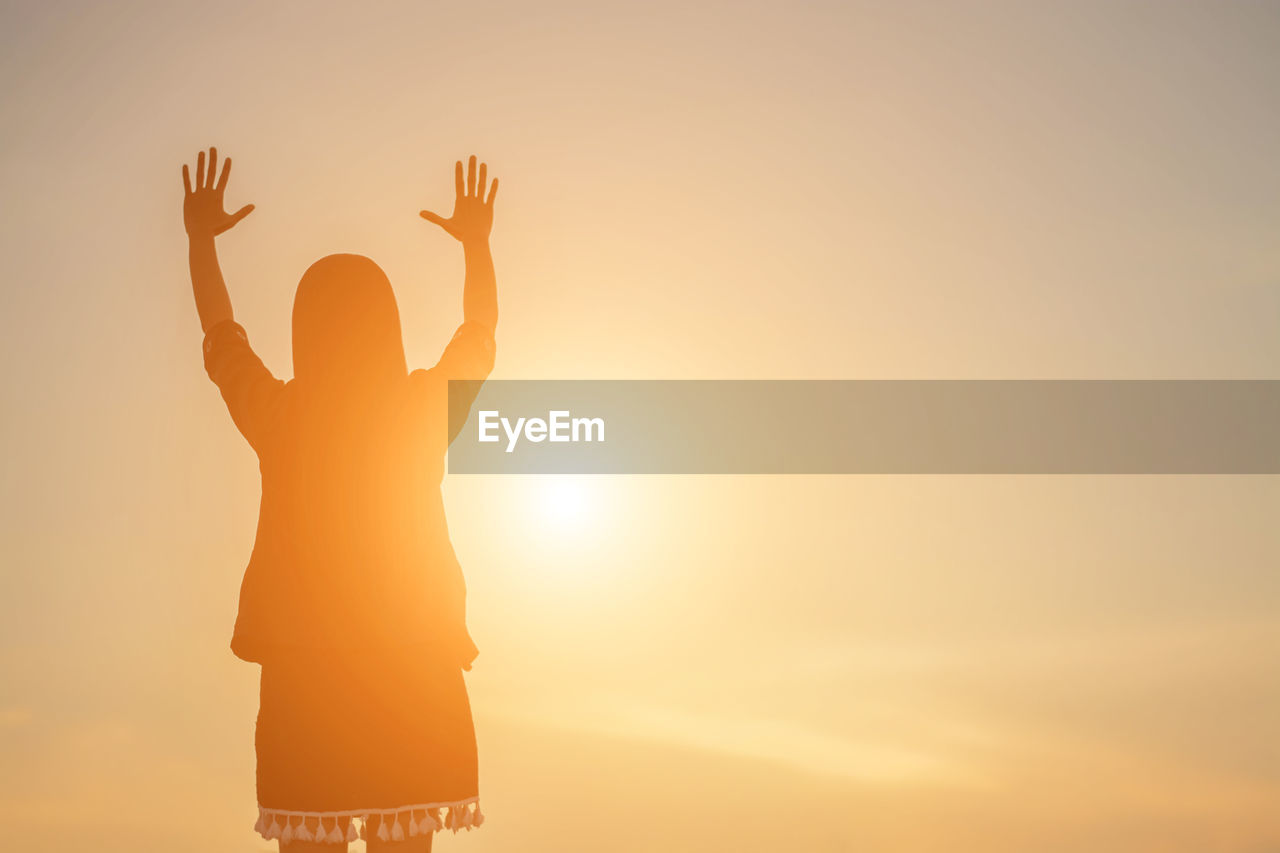 Rear view of silhouette woman with arms raised standing against sky during sunrise