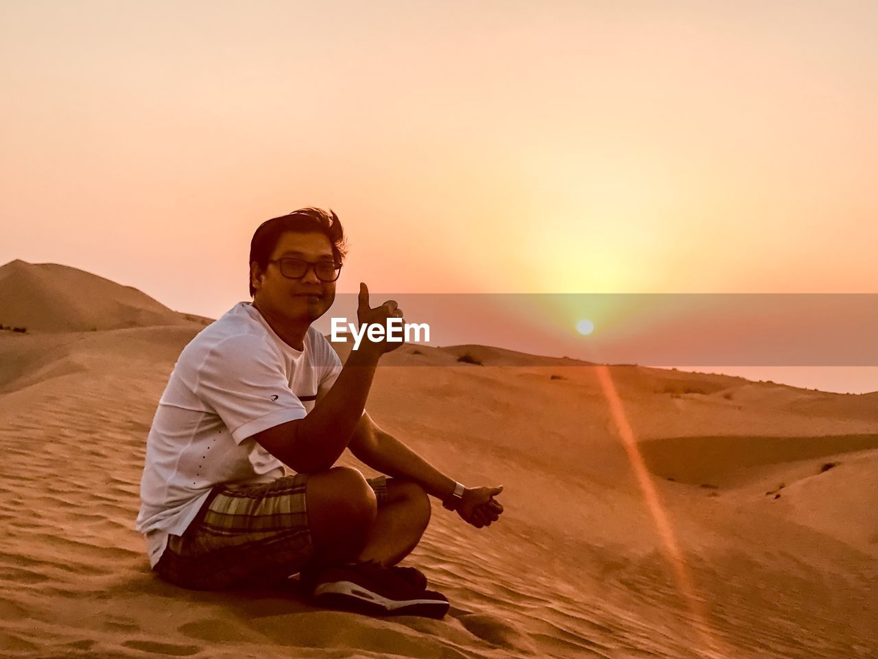 FULL LENGTH PORTRAIT OF YOUNG MAN SITTING ON SAND AT SUNSET