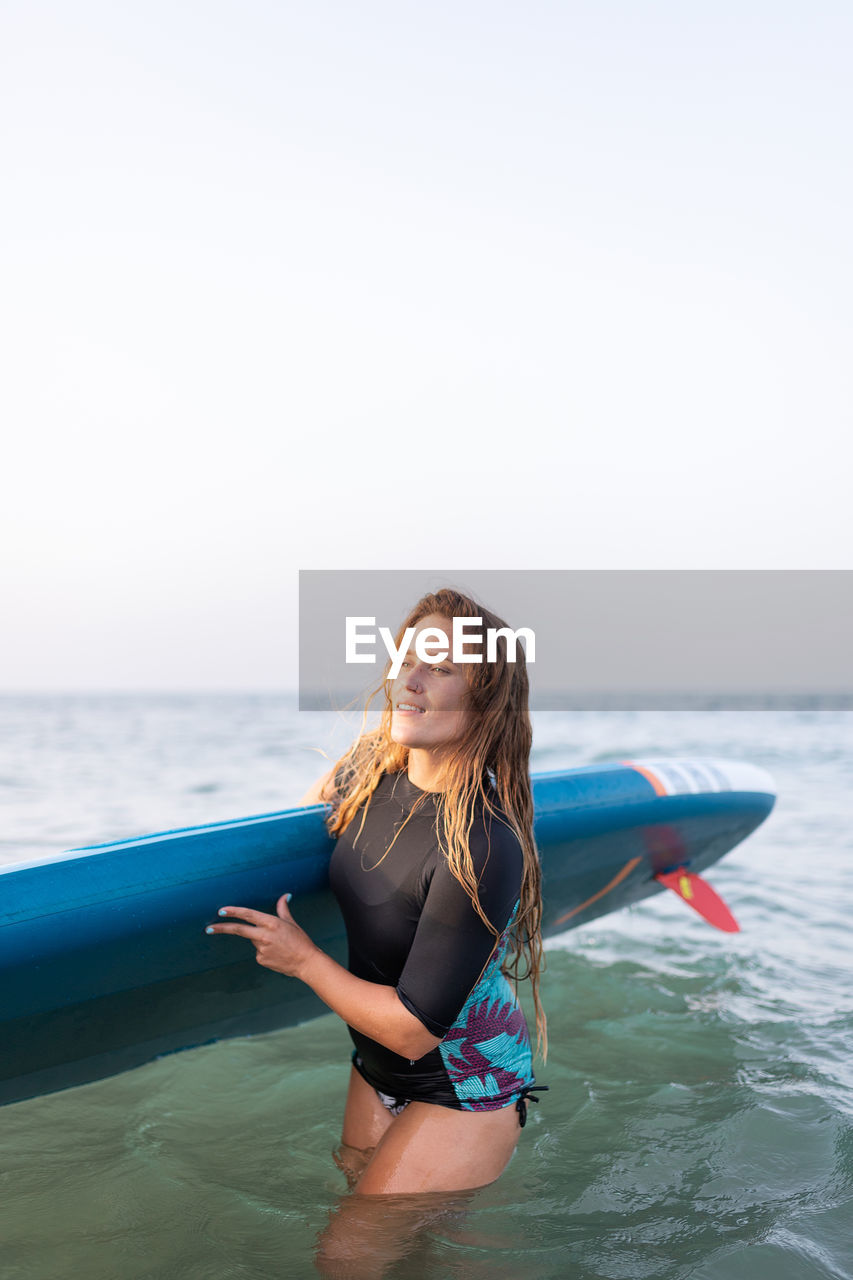 Female in swimsuit standing with sup board in sea water in summer and looking away