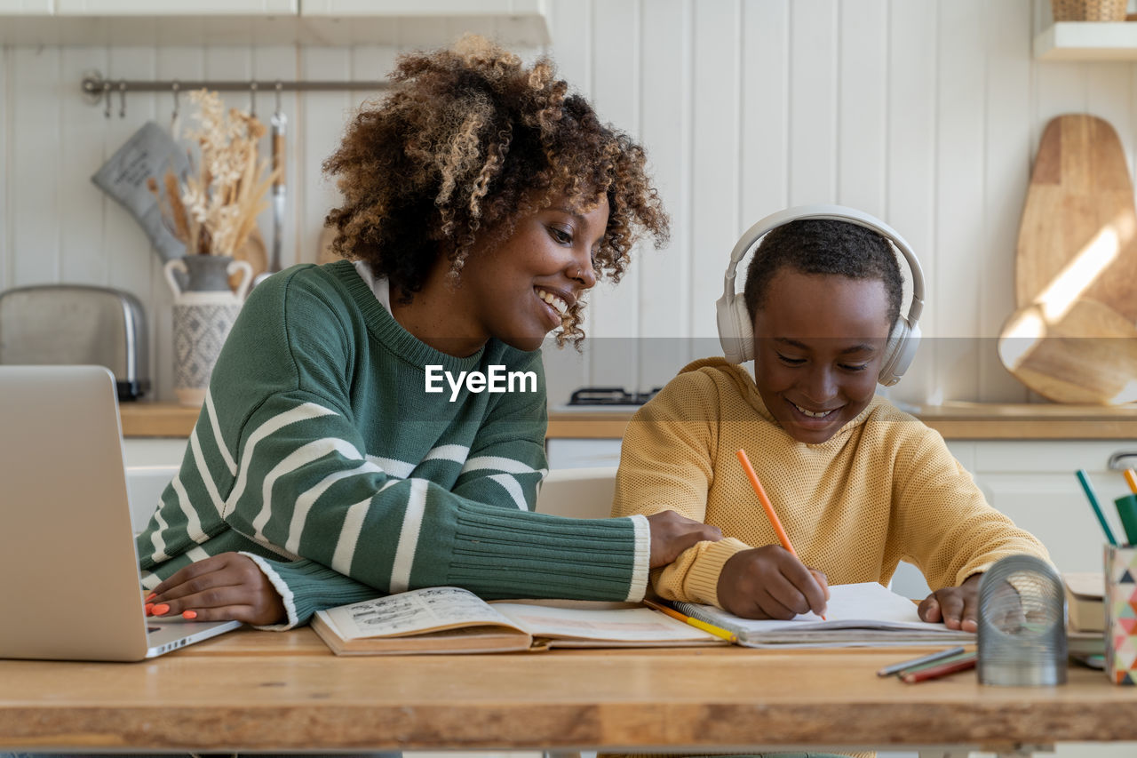 Happy smiled black child boy with pleased mother sit at desk with pencil write at school tutorial