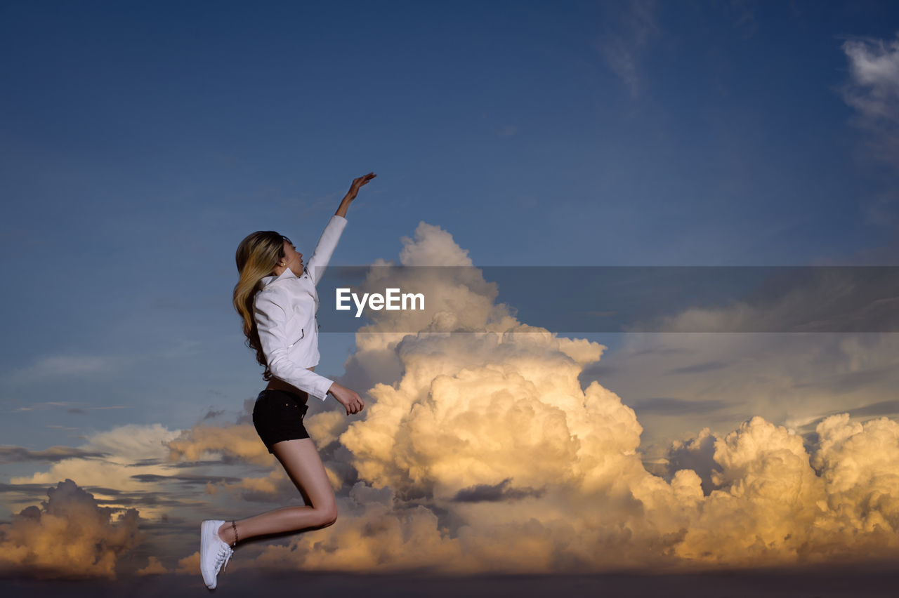 Side view of young woman jumping against cloudy sky