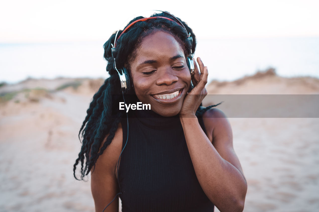 Optimistic african american female in headphones enjoying songs and dancing with closed eyes on sandy shore against sea in summer evening