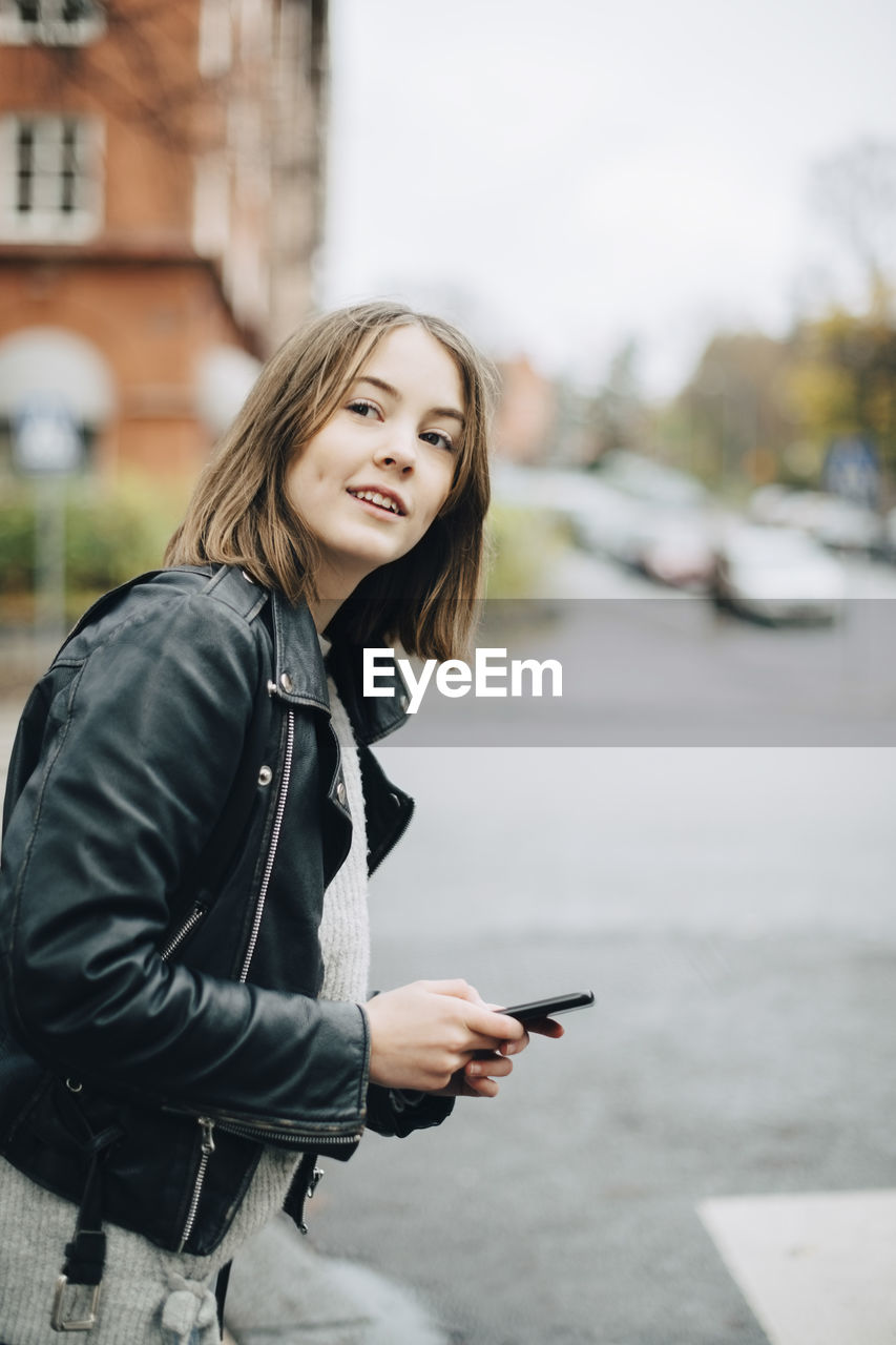 Smiling girl with mobile phone looking away while standing on street in city