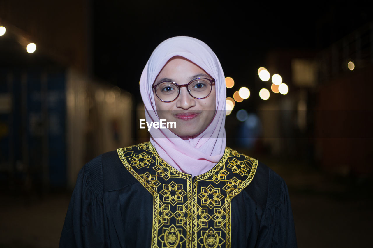 Smiling young woman wearing mortar board while standing against sky at night