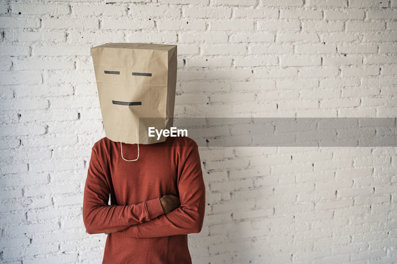 Man with arms crossed wearing brown paper bag on face against white brick wall