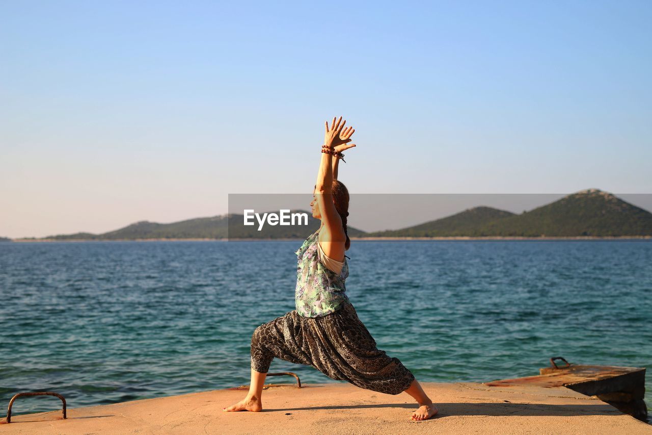 Woman with arms raised doing yoga at beach against clear sky
