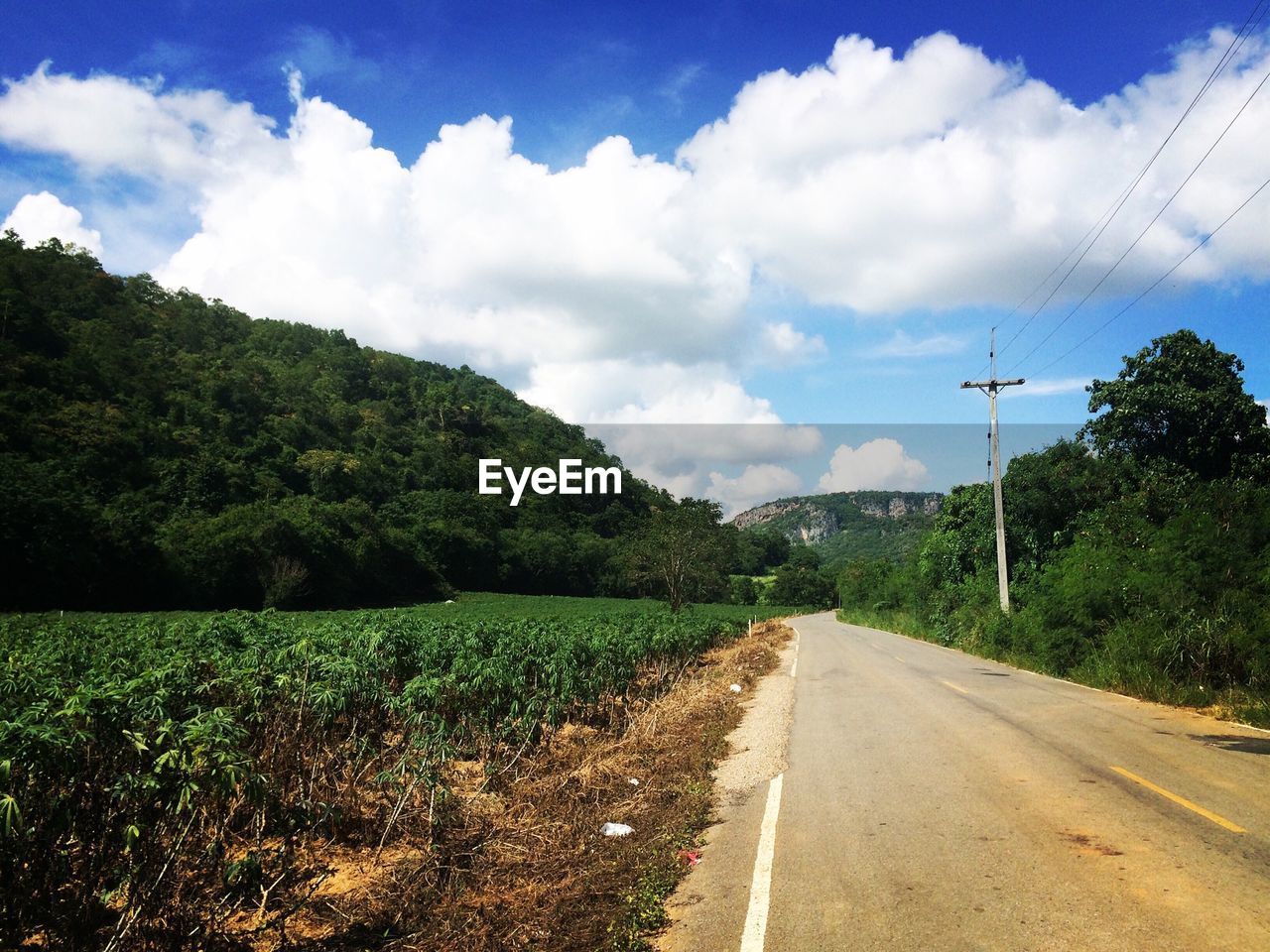 Empty road by trees leading towards mountain against sky