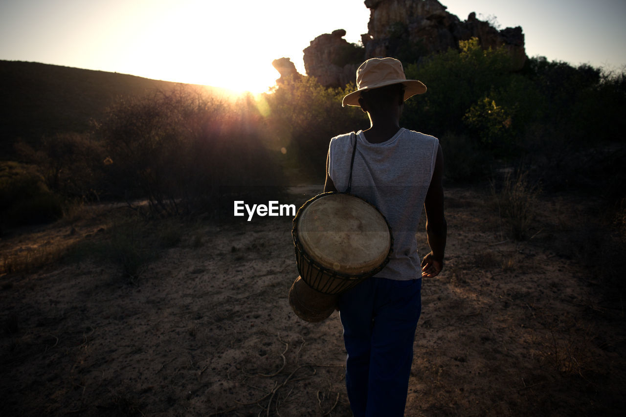 Rear view of man with musical instrument standing against trees during sunset