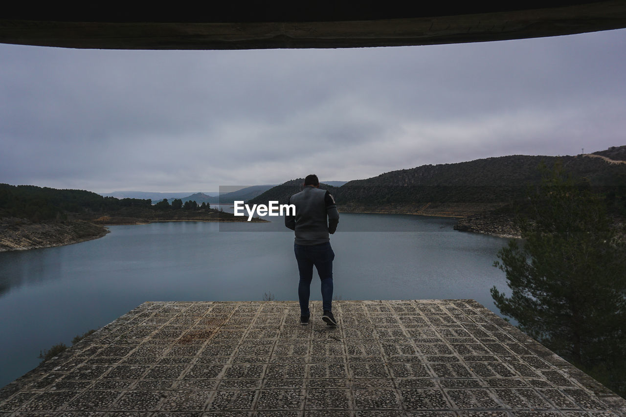 Rear view of man walking at observation point by river against cloudy sky