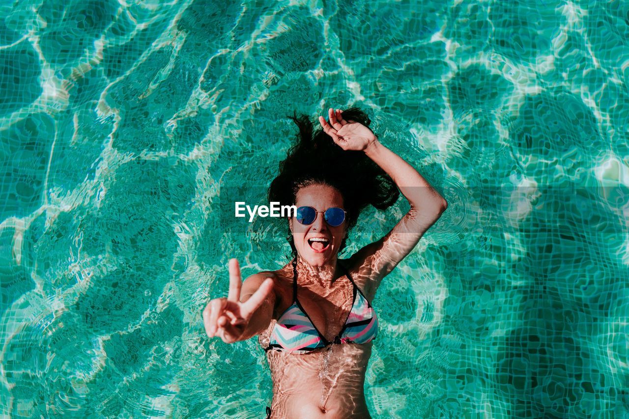 Portrait of cheerful woman showing peace sign while swimming in pool