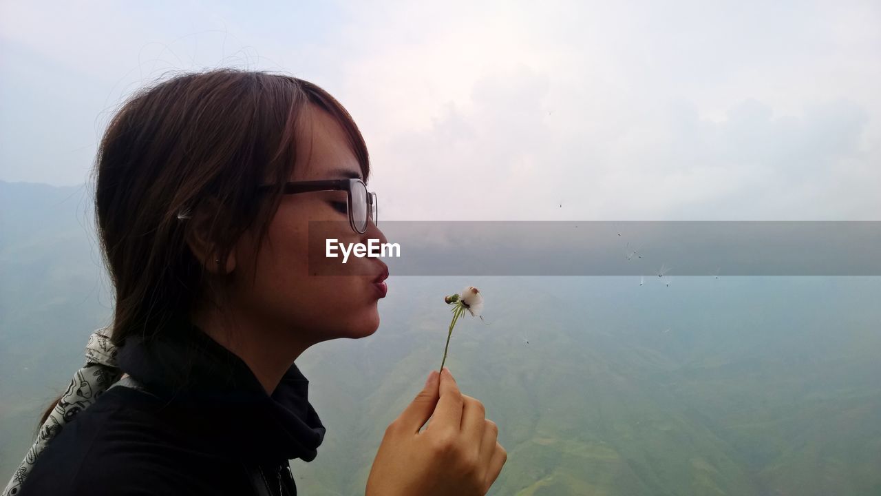 Close-up of young woman blowing dandelion seeds against mountains