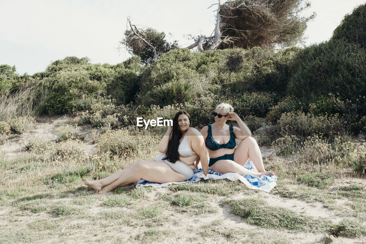 Delighted curvy friendly females in bikini sitting on blanket on shore and enjoying sunny day in summer