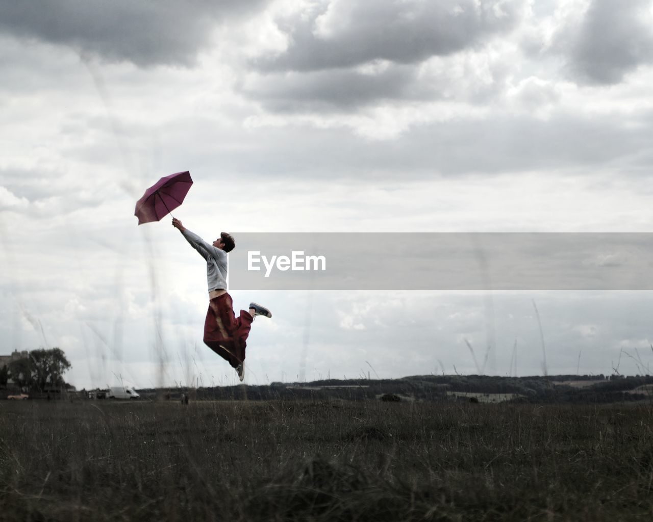 Side view of young man with umbrella jumping on grassy field against cloudy sky