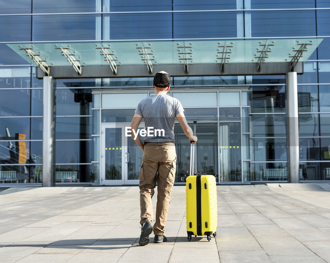 Rear view of young man traveler in casual carrying yellow suitcase next to entrance to airport 