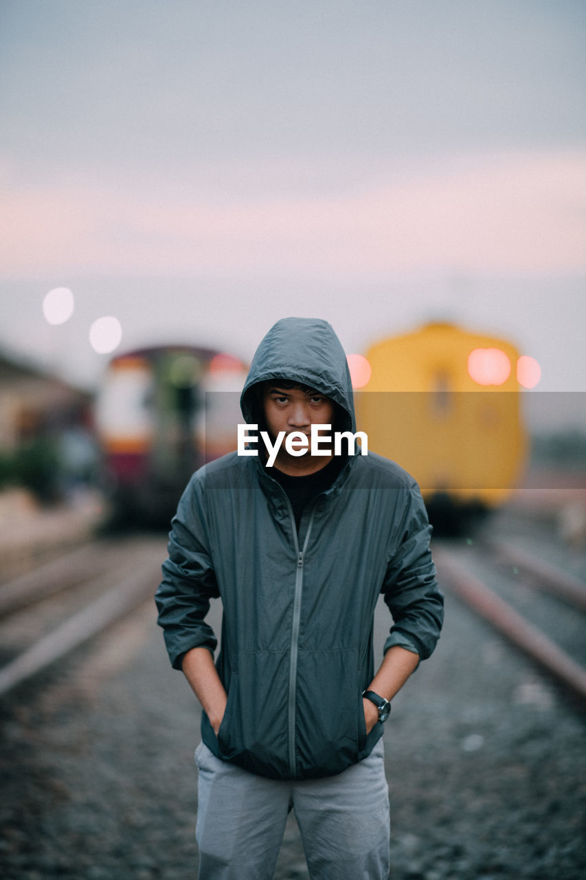 Portrait of man wearing hooded jacket while standing against trains at railroad tracks